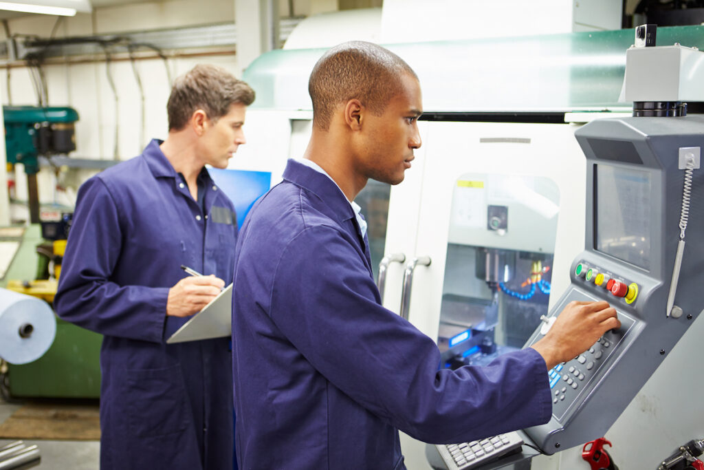 Engineer And Apprentice Using Automated Milling Machine At Work