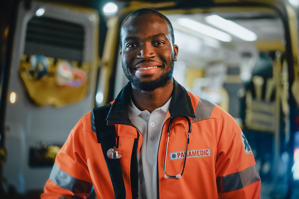 Portrait of a Black African American EMS Paramedic Proudly Standing in Front of Camera in High Visibility Medical Orange Uniform and Smiling. Successful Emergency Medical Technician or Doctor at Work.